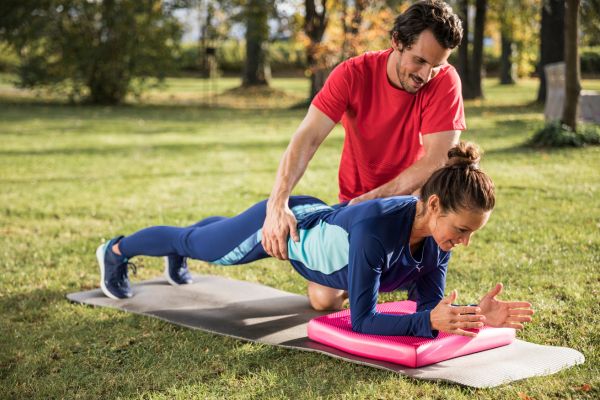 Eine Frau führt einen Plank zur Stärkung der Tiefenmuskulatur aus und ein Trainer unterstützt sie.